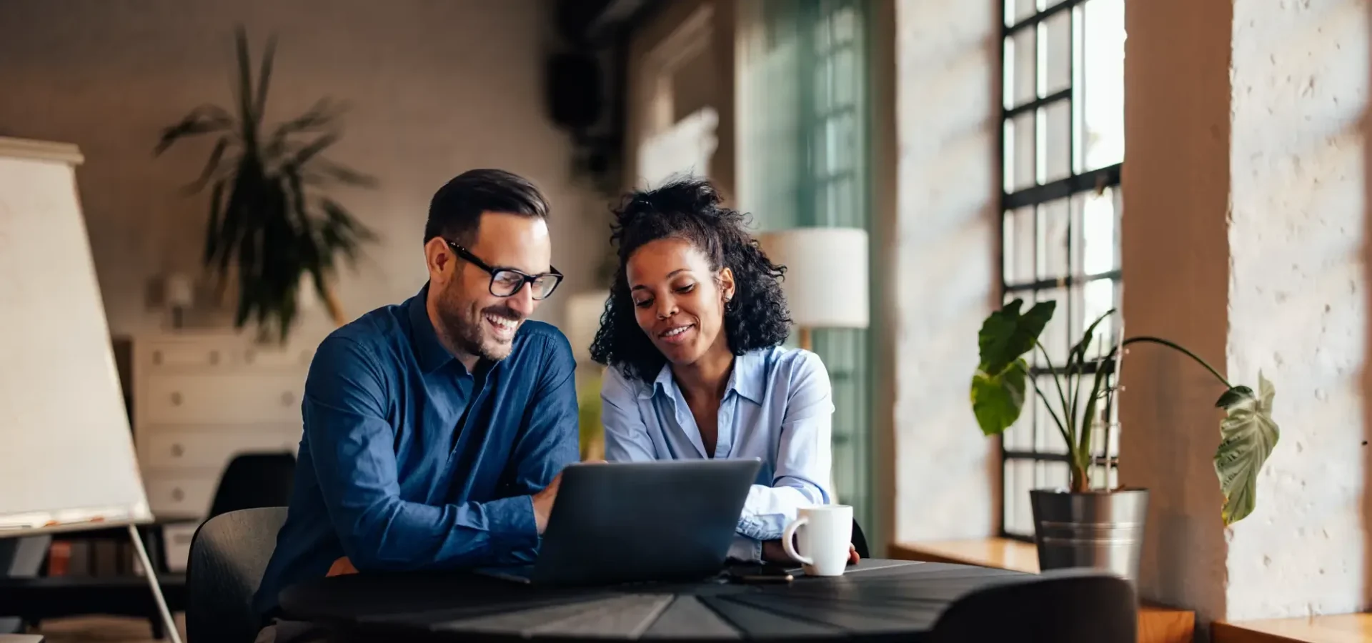 Two professionals collaborating at a laptop in a modern office with natural light and plants.