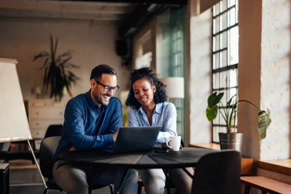 Two professionals collaborating at a laptop in a modern office with natural light and plants.