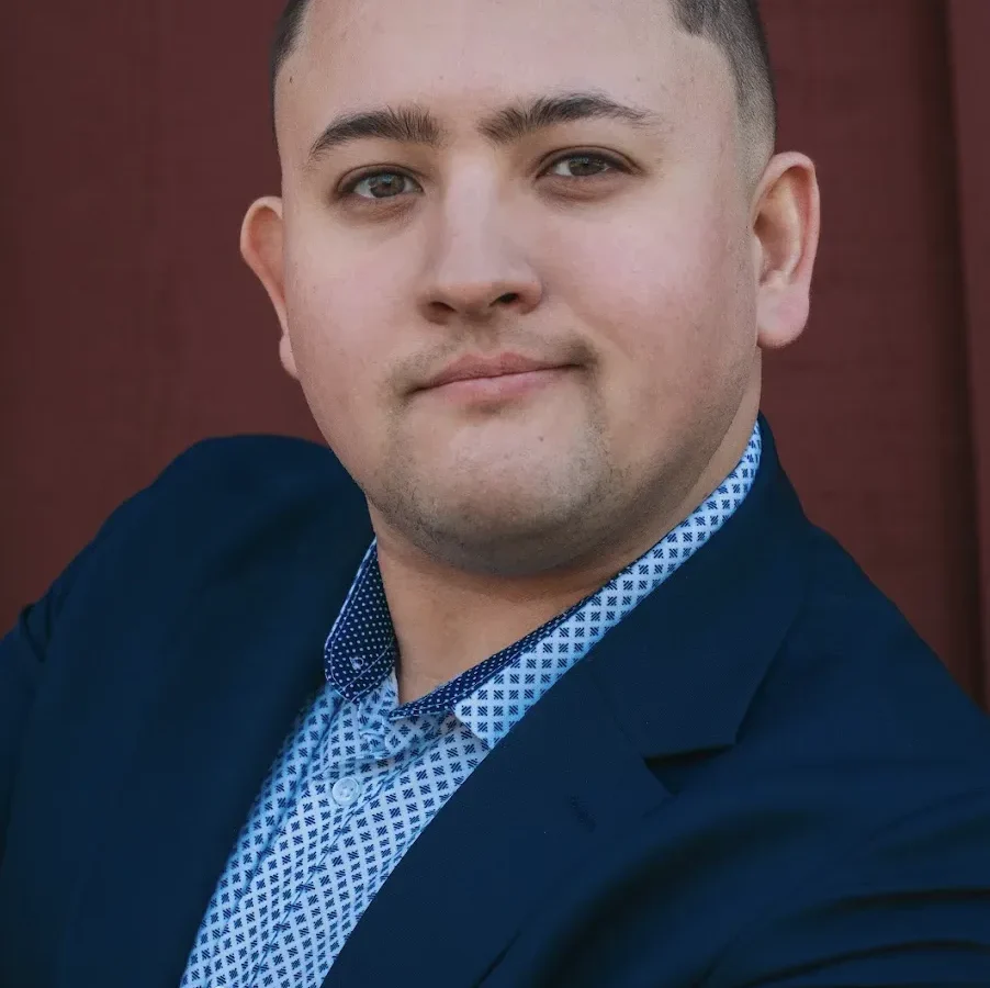Professional headshot of a man in a dark blue blazer and patterned blue shirt against a burgundy background.