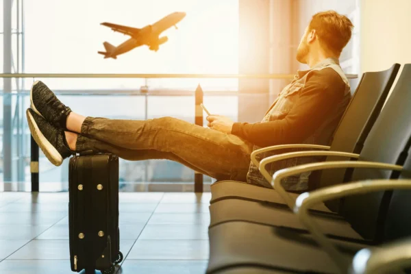 Man relaxing in airport terminal with feet on luggage, watching airplane take off through window.