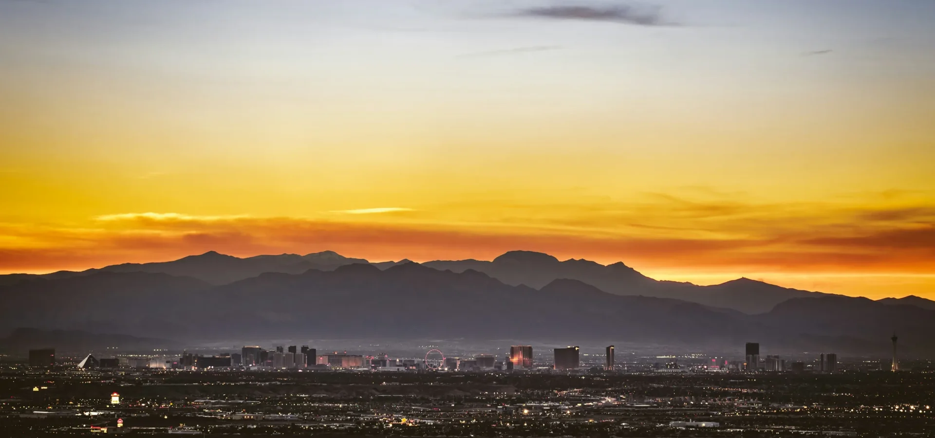 Sunset view of Las Vegas skyline with mountains silhouetted against golden and orange sky, city lights beginning to glow.